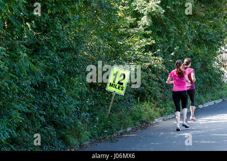 Zwei junge, weibliche / Sportlerinnen Sport Kleidung tragen im Bild laufen vorbei an eine 12-Meilen-Marker-Schild an einem Halbmarathon-Straßenrennen in Großbritannien Stockfoto