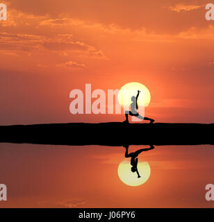 Silhouette einer ständigen sportliche Frau Yoga zu praktizieren mit auferweckt Arme auf dem Hügel nahe dem See mit Himmel Spiegelung im Wasser. Sonne und Orange Himmel w Stockfoto