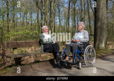 Ältere Frau im Rollstuhl im Gespräch mit ihr ehemaliger Ehemann sitzt auf einer Parkbank beim Spaziergang im Wald an einem sonnigen Tag im Frühjahr deaktiviert Stockfoto