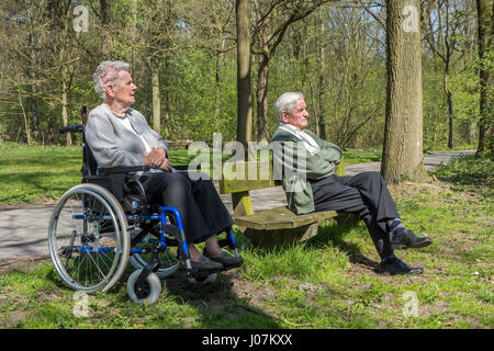 Behinderte ältere Frau im Rollstuhl und pensionierte Mann sitzt auf einer Parkbank, die Natur beim Spaziergang im Wald an einem sonnigen Tag im Frühling genießen Stockfoto