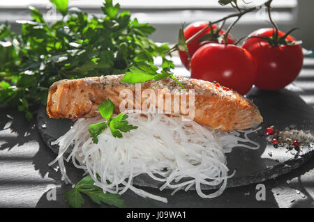 Cellophan mit Stücken von gebratener Lachs mit Tomaten, Zwiebeln und Petersilie auf dem Brett von schwarzem Schiefer Stockfoto