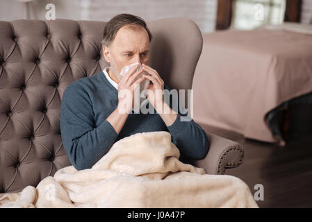 Krank zu werden. Senior Mann sitzt auf Couch zu Hause mit warmen Decke bedeckt und seine Nase weht. Stockfoto