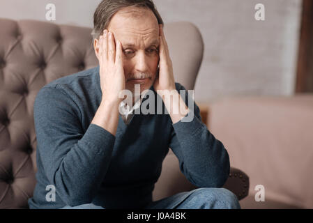 Was passiert ist. Nahaufnahme von frustrierten senior Mann auf der Couch sitzen und seine Schläfen mit Händen zu berühren. Stockfoto