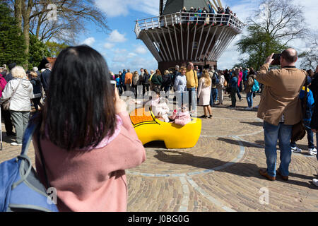 Zwei asiatische Mädchen posiert in einem riesigen hölzernen Schuh mit einer Windmühle im Hintergrund, während ihre Mutter ein Foto mit ihrem Handy - Frühling 2017 in Keuke nimmt Stockfoto