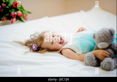 Das charmante kleine Mädchen liegt auf einem großen Bett. In Händen auf das Baby ein grauen Teddybär. Das Mädchen hat einen fröhlich verspielte Look und das Haar zerzaust aus einem p Stockfoto