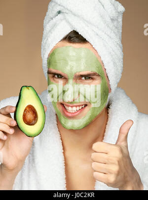 Mann trägt Avocado Gesichtsmaske und Daumen nach oben zeigen. Porträt von glücklicher Mann mit Gesichtsmaske und Holding Scheibe Avocado. Schönheit & Skin care Konzept Stockfoto