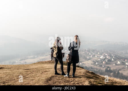 Zwei junge nette polnische Mädchen (einer von ihnen Telefonieren) mit langen braunen Haaren auf ochodzita Hügel Gipfel mit koniakow Dorf unten im späten Herbst schlesischen b Stockfoto