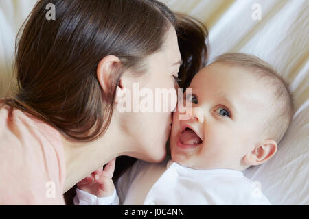 Porträt von niedlichen Babymädchen wird von Mutter auf Bett geküsst Stockfoto