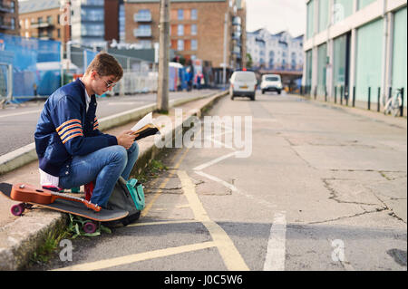 Junger Mann sitzt auf Kerb, Buch, Skateboard neben ihm, Bristol, UK Stockfoto