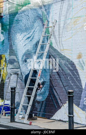 Ein Künstler, trug eine schwarze Kapuzenjacke schafft ein Kunstwerk durch das Malen ein Wandbild an der Wand in der Hanbury Street in East London. Stockfoto