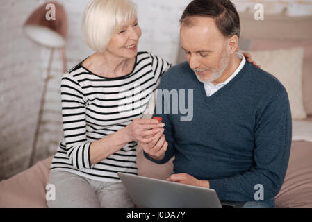 Mit großer Liebe. Lächelnd senior Frau ist an ihren Mann mit Laptop auf dem Bett sitzend ein Heilmittel geben. Stockfoto