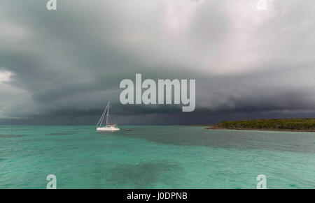 Eine dunkle Bö Gewitterwolke über das ruhige grüne Wasser einer Bahama Bucht als ein Katamaran Segelboot liegt in einer ruhigen Ankerplatz. Stockfoto