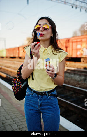 Junge Teenager-Mädchen stehen auf der Plattform am Bahnhof und bläst Seife Luftblasen, am gelben T-shirt, Jeans und Sonnenbrille mit Rucksack zu tragen Stockfoto