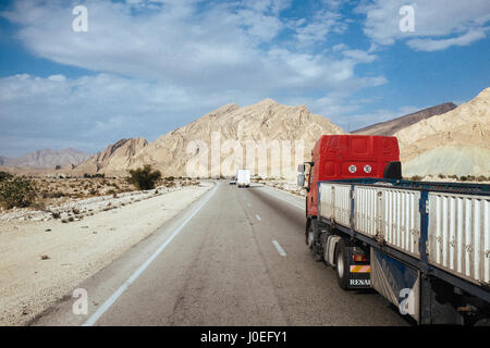 LKW sind durch die beeindruckende Landschaft mit Bergen, blauem Himmel und weißen Wolken auf der Fahrbahn zwischen Bandar Abbas und Sirjan in Fahrt. Stockfoto