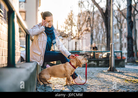 Frau mit einem Hund reden auf Smart Phone und sitzt auf einem grünen Metallzaun Stockfoto