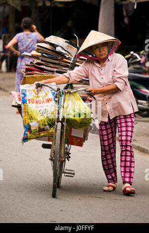 Vertikale Porträt einer alten Dame schob ein Fahrrad beladen mit Kartons durch die Straßen in Hoi an, Vietnam. Stockfoto