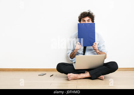 Ein barfuß Geschäftsmann auf dem Boden an einer Wand sitzen und einen Laptop verwenden und bedeckte sein Gesicht mit flipchart Stockfoto