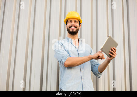 Arbeiter mit gelben Helm und Jeans Hemd in der Nähe einer industriellen Wand mit einer Tablette um zu sehen, das Projekt Stockfoto