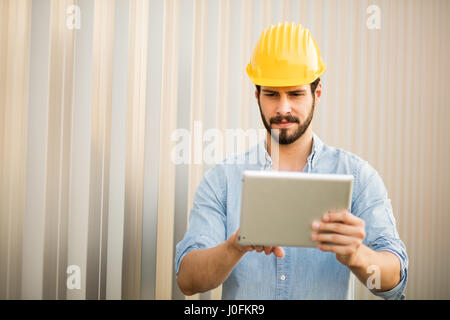 Arbeiter mit gelben Helm und Jeans Hemd in der Nähe einer industriellen Wand mit einer Tablette um zu sehen, das Projekt Stockfoto