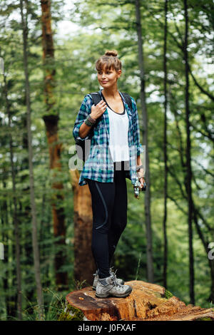 alleinreisende Frau sitzt auf einem Baumstamm mit ihrem Rucksack und Kamera mitten im Wald beobachten und die Natur genießen Stockfoto