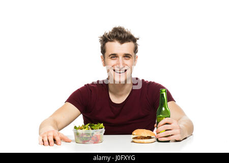 junger Mann mit vor zwei Möglichkeiten, Bier und Fast Food vs. Veganer Salat, am Tisch isoliert auf weiss Stockfoto