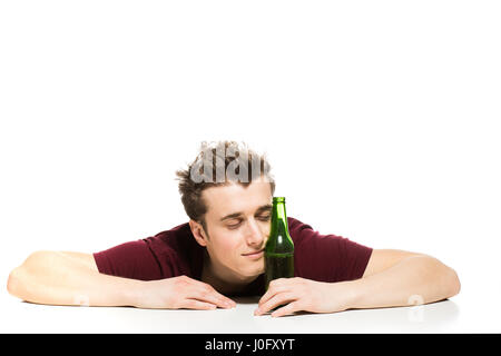 verschlafene junger Mann mit einer Flasche Bier am Tisch, isoliert auf weiss Stockfoto