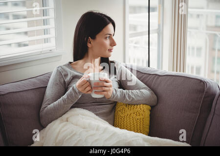 Frau, die Tasse beim Blick durch Fenster zu Hause Stockfoto