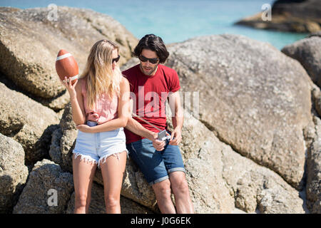 Junges Paar beim Sitzen auf Felsformation am Strand im sonnigen Tag im Gespräch Stockfoto