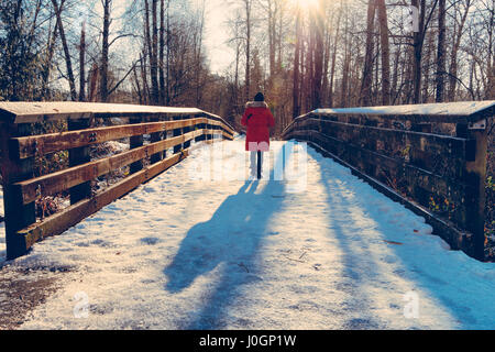 Frau im roten Mantel werfen lange Schatten über Schnee bedeckte Brücke im Wald wandern. Stockfoto
