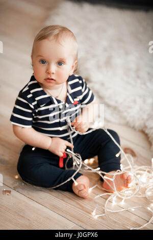 Kleines Baby sitzt auf Holzboden und spielt mit Kranz von Glühbirnen. Er schaut ernst. Stockfoto