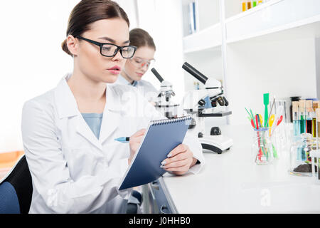 Junge Wissenschaftlerin, die Notizen in der Nähe von Kollegin arbeiten mit Mikroskop Stockfoto