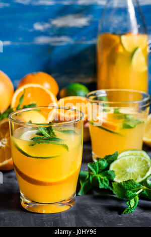 Erfrischende Getränke, Zitrusfrüchte, Orangen Limonade Kalk frischen Minze in Flasche und Gläser, geschnittene Früchte auf Holztisch, blauer Hintergrund, Fenster Stockfoto