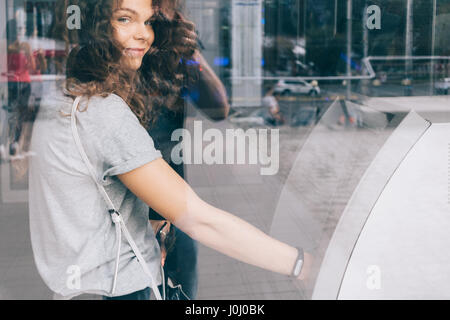 Lockige lächelndes Mädchen zieht Geld von einem Geldautomaten in der Stadt, die Reflexion von der Straße auf dem Glas Stockfoto