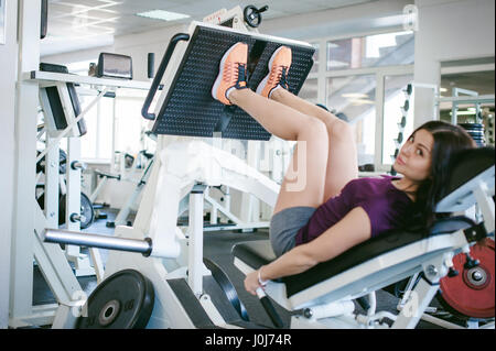 Mädchen in einem hellen weißen Fitnessstudio. Eine Brünette Frau eine macht Übung für eine Gruppe von Beinmuskulatur auf einem Simulator zu tun. Gesunde Lebensweise, gute Laune Stockfoto