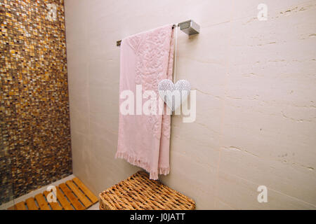 Ein rosa Handtuch im Bad auf der beheizten Handtuchhalter getrocknet. Weißes Herz, ein Element des Dekors. Weidenkorb für Kleidung. Stockfoto