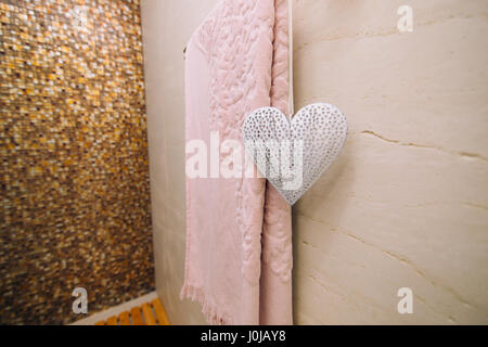 Ein rosa Handtuch im Bad auf der beheizten Handtuchhalter getrocknet. Weißes Herz, ein Element des Dekors. Weidenkorb für Kleidung. Stockfoto