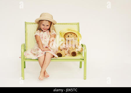 Kleines Mädchen, Kinder tragen ein Sommerkleidchen und Hut mit passenden Teddybär sitzen zwei zweiersitzbank mit einem schüchternen Blick auf ihrem Gesicht Stockfoto