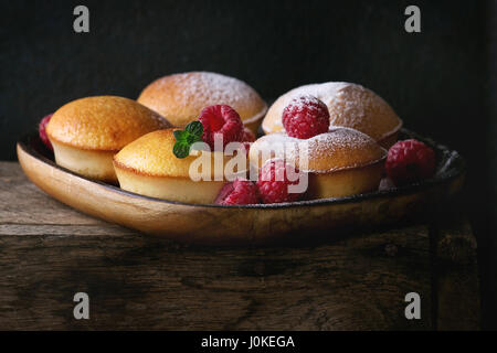 Hausgemachte Zitronen Muffin Muffins mit frischen Himbeeren, Zucker Pulver, Minze, in quadratische Holzplatte auf Holztisch auf dunklem Hintergrund serviert. Dunklen Rost Stockfoto