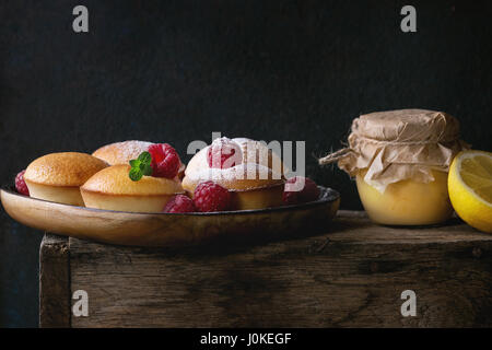 Hausgemachte Zitronen Muffin Muffins mit frischen Himbeeren, Zucker Pulver, Minze, serviert mit Glas Lemon Curd und Zitronen auf Holzkiste auf dunklem Hintergrund Stockfoto