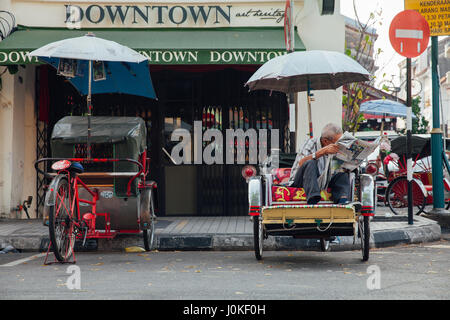 George Town, Deutschland - 21. März 2016: Rikscha Zeitunglesen sitzen auf dem Dreirad an der Straße von George Town, Penang, Malaysia auf Ma Stockfoto