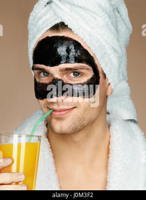 Glücklicher Mann mit schwarzer Maske auf dem Gesicht Getränke Saft. Foto von gepflegten Mann Spa-Behandlungen erhalten. Schönheit & Skin care Konzept Stockfoto