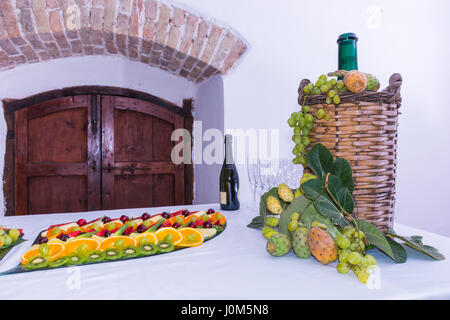Zusammensetzung der Früchte und geschliffenem Glas Korbflasche beschichtet für Wein Wicker. Auf einem Tisch, beladen mit einem Restaurant, mit Trauben, Feigen, Kaktusfeigen und Blätter. A Stockfoto