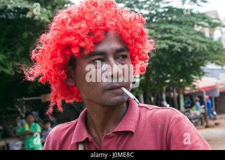 Ein kambodschanischer Mann raucht eine Zigarette während des Tragens einer roten Perücke als er Feierlichkeiten Khmer Neujahr in Chork Dorf, Tboung Provinz, Kambodscha. Stockfoto