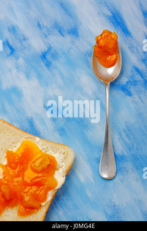 Toastscheibe mit Butter und Marmelade und Silberlöffel mit Marmelade Blob über blau bemalte Textilien Hintergrund. Draufsicht. Stockfoto