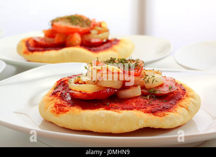 Kleine Pizza (Pizzette) auf Teller mit Wurst, Zwiebeln und Tomaten Scheibe. Unkonzentriert zweite Pizza auf Hintergrund. Stockfoto