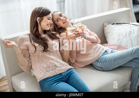 Zwei attraktive junge Frauen sitzen auf dem Sofa und jubeln mit Weißwein Stockfoto