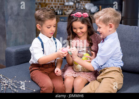 Zwei kleine Jungen und Mädchen auf dem Sofa sitzen und halten Ostereier Stockfoto