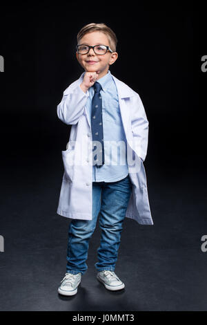 Kleiner junge Wissenschaftler in Brillen und Lab Coat Holding hand aufs Kinn und lächelnd Stockfoto