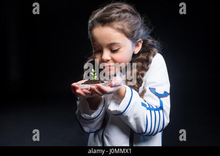 Niedliche kleine Mädchen in Astronaut Kostüm mit frischen Pflanze im Boden auf schwarz Stockfoto