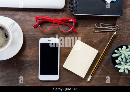 Draufsicht der Kaffeetasse, Smartphone mit leerer Bildschirm, Notebook, rote Brille und Sukkulente im Topf auf Holztisch Stockfoto